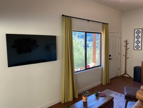 Un salón con un ventanal grande, un televisor de pantalla plana montado en la pared, una mesa de centro de madera y una puerta con perchero.