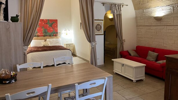 A room with a bed, a red sofa, a dining table with chairs, and a small white table. - SUITE DI SAN FRANCESCO (Grottaglie)
