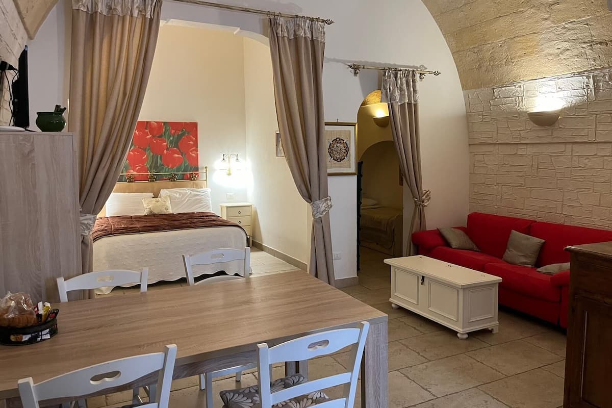 Una habitación con una cama, un sofá rojo, una mesa de comedor con sillas y una pequeña mesa blanca.