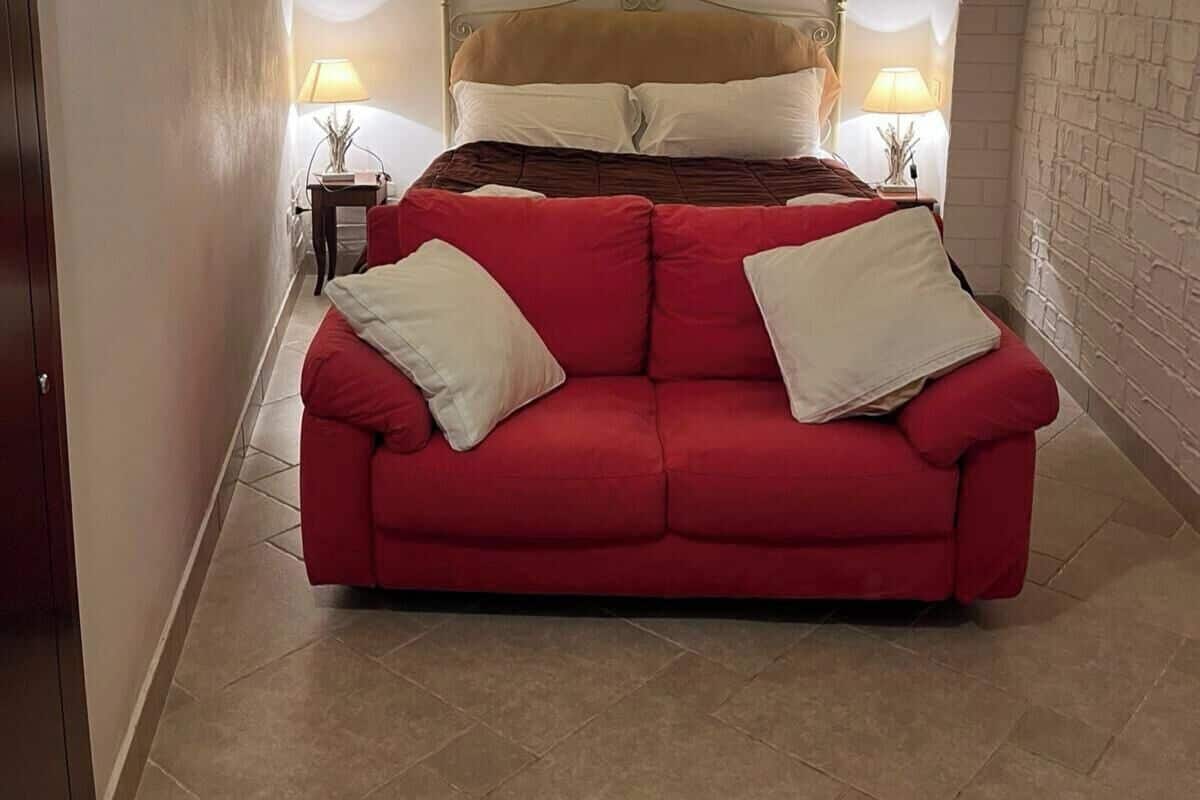 Un sofá rojo con almohadillas blancas en una habitación que incluye una cama, dos mesitas de noche con lámparas y un suelo de baldosas.