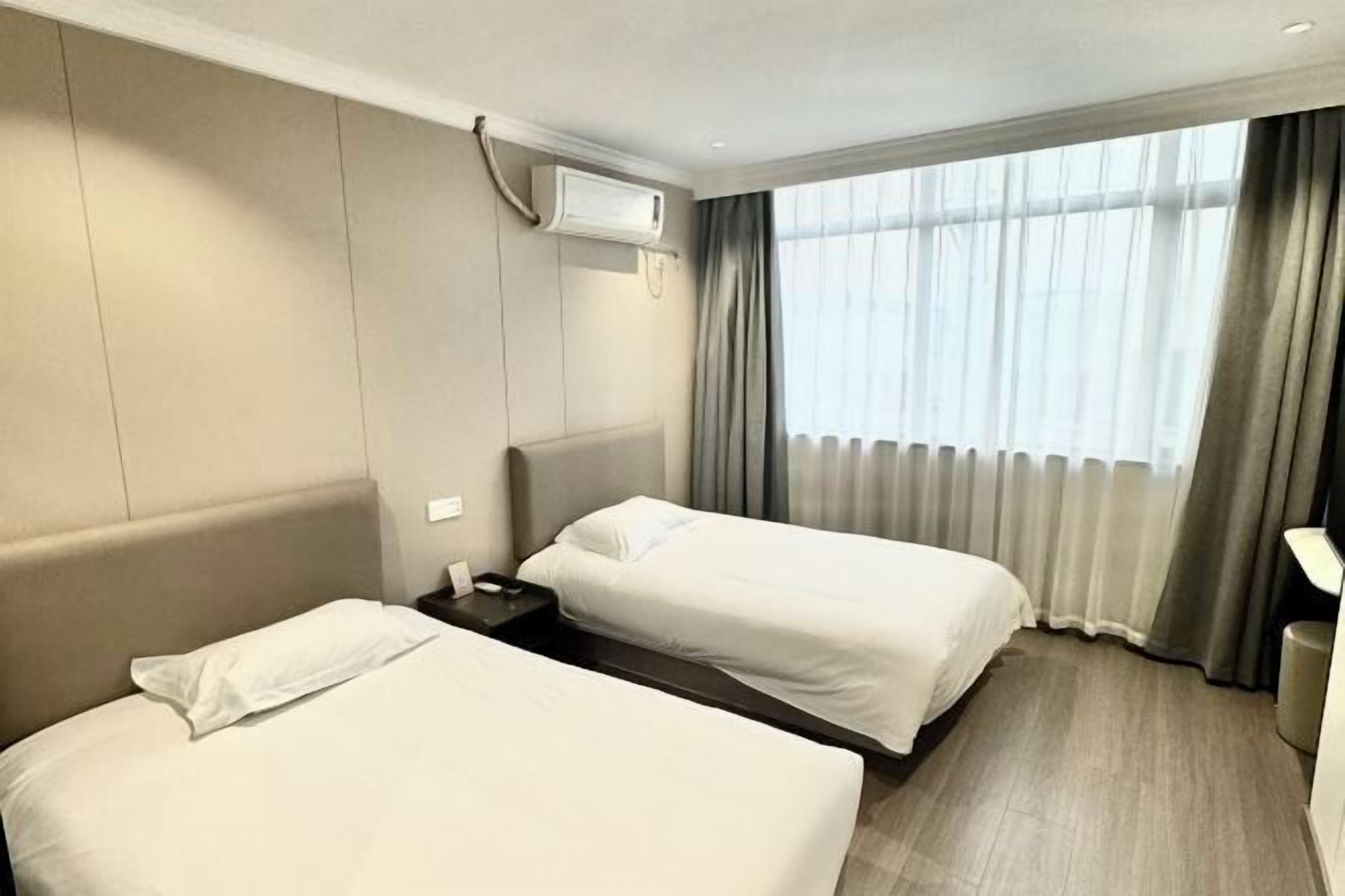 A hotel room with two beds, a bedside table, an air conditioning unit, and a window with curtains.