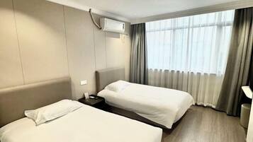 A hotel room with two beds, a bedside table, an air conditioning unit, and a window with curtains.