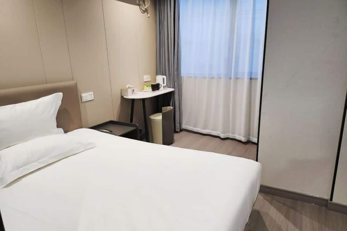 Une chambre d’hôtel avec un lit, une petite table sur laquelle se trouvent une tasse et une poubelle, une fenêtre avec des rideaux et un luminaire fixé au mur.