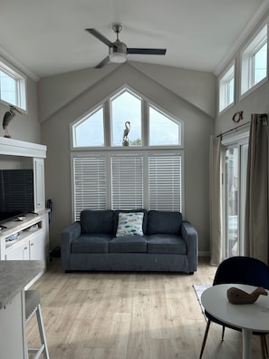 A modern living room with a grey sofa, a ceiling fan, and a large window with blinds. - Keys Paradise! (Key Largo)