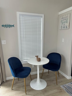 A room with a white round table, two blue chairs, a white window with blinds, a wall decoration, and a small decorative item on the table. - Keys Paradise! (Key Largo)