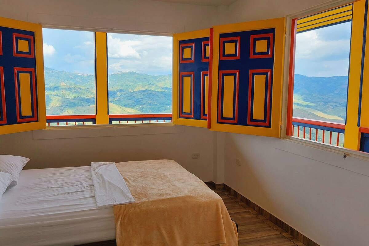 Ein Zimmer mit einem Bett, einem Fenster mit Bergblick und bunten Fensterläden.