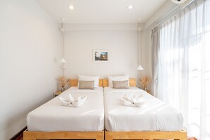 Ein Doppelzimmer mit zwei Betten, jeweils mit weißer Bettwäsche und dekorativen Kissen, einem kleinen gerahmten Bild an der Wand und einem großen Fenster mit durchsichtigen Vorhängen.