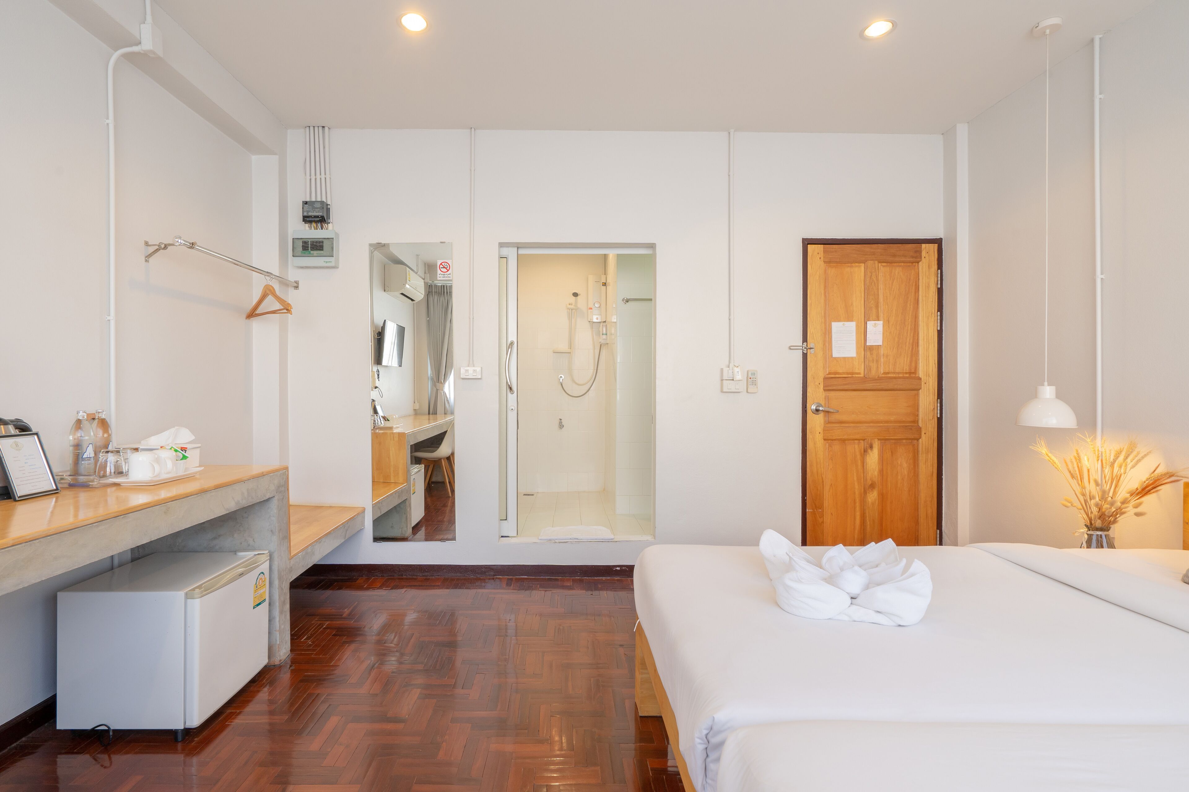 Habitació d'hotel amb terra de fusta, llit amb roba de llit blanca, escriptori amb un ordinador, nevera petita, porta de fusta i zona de dutxa.