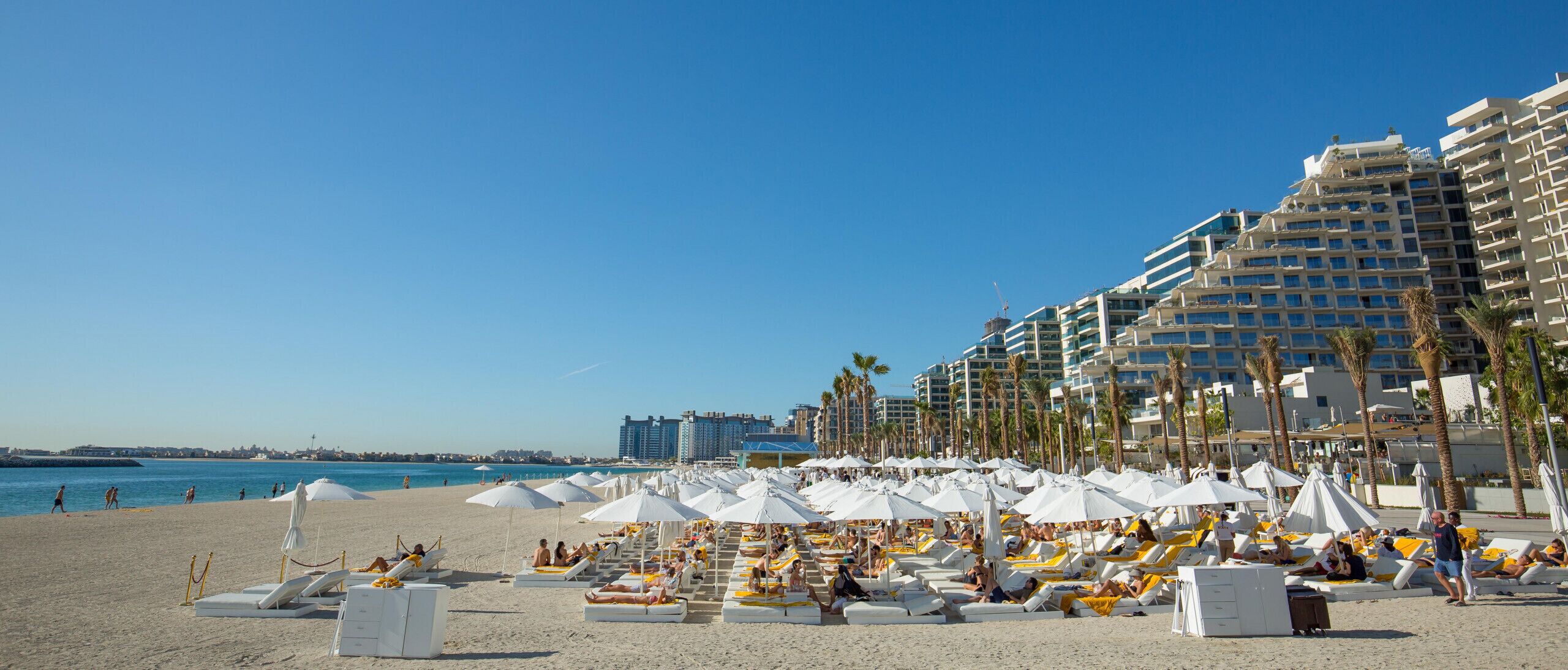 Una platja amb gandules i ombrelles, cel blau i un hotel al fons.