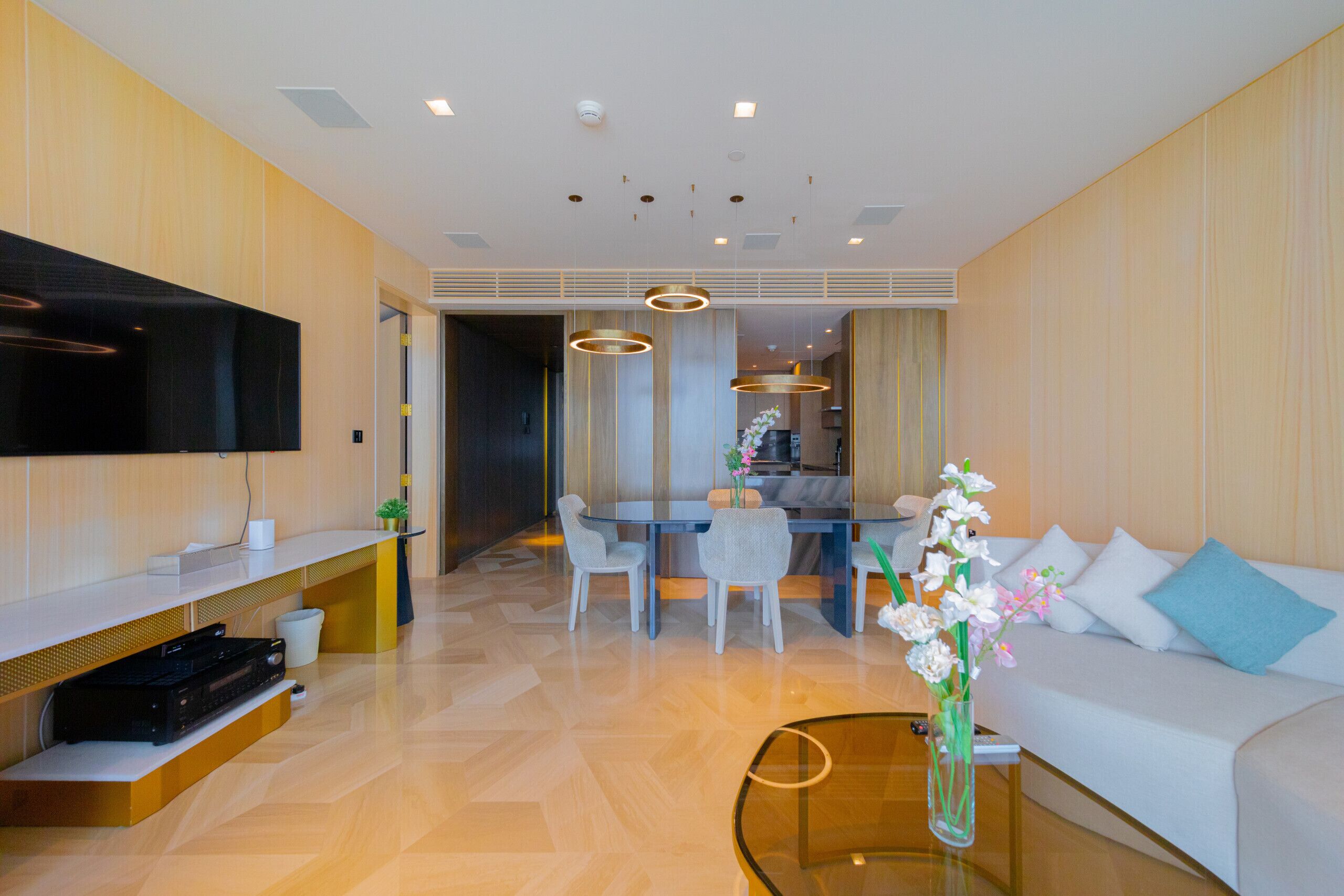 A modern hotel room with a flat-screen TV, a dining area with chairs and a table, a sofa with cushions, and a glass coffee table with a vase of flowers.
