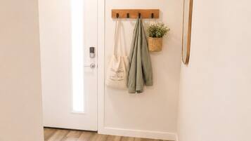 Un couloir avec un plancher en bois, un porte-manteau avec des sacs et une plante, une porte avec un interrupteur et un miroir accroché au mur.