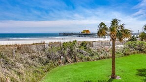 A beach view with a pier, a grassy area, and a palm tree. - Margate Penthouse Myrtle Beach (Myrtle Beach)