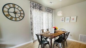 A dining area with a wooden table and chairs, a wall clock, and a window with curtains. - Live like a local in our cozy, clean home! (Fayetteville)