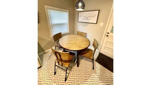 A dining area with a round wooden table and four chairs, a pendant light, and a framed sign on the wall.