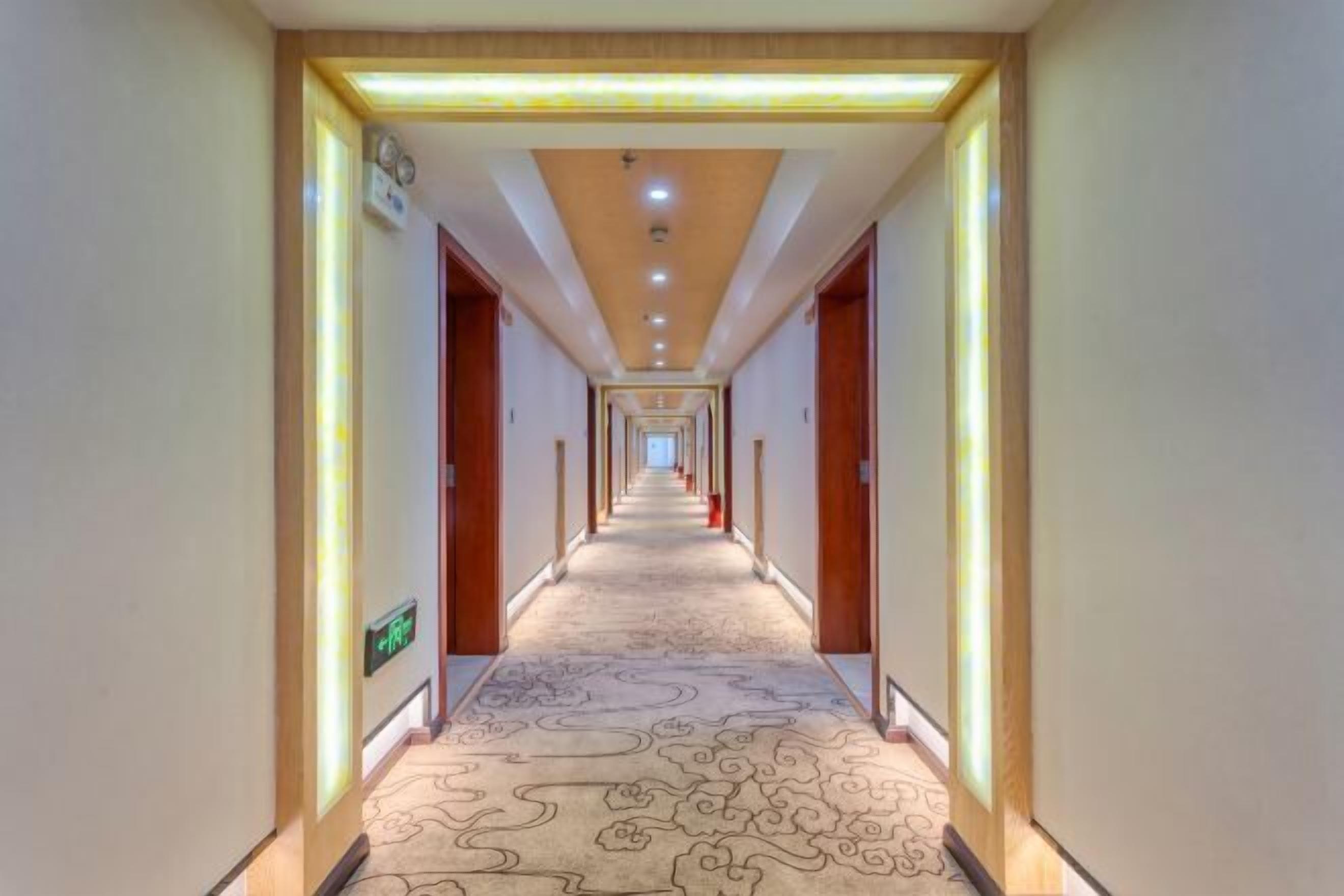 Un largo y estrecho pasillo de hotel con alfombra estampada, puertas de madera e iluminación empotrada en el techo.