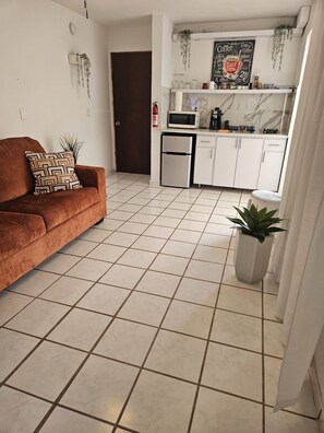 A living room with a sofa and a coffee table, a kitchen with white cabinets and a microwave, and a tiled floor.