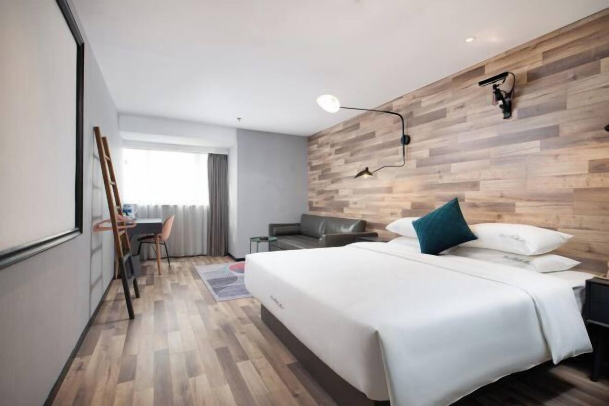 Ein modernes Hotelzimmer mit einer Holzwand, einem Bett mit weißer Bettwäsche, einem Schreibtisch mit Stuhl und einem Fenster mit Vorhängen.