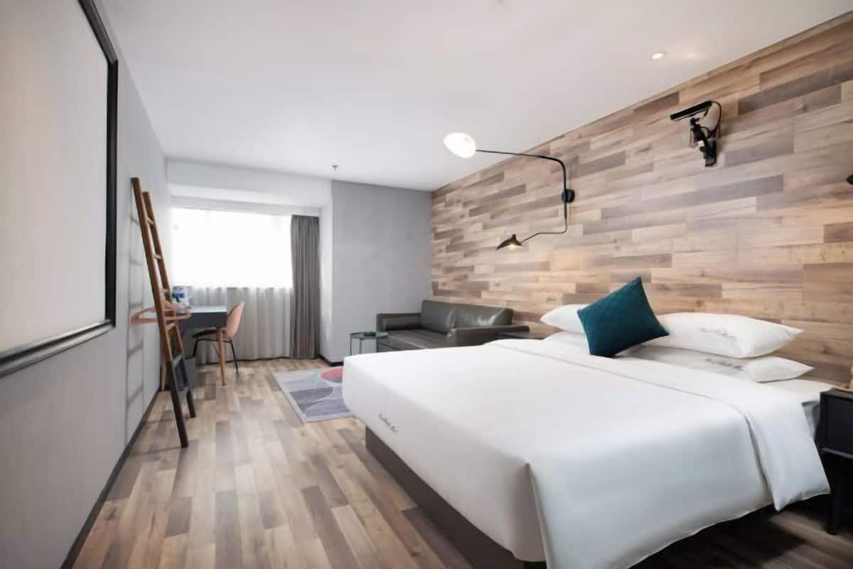 A modern hotel room with a wooden wall, a bed with white bedding, a desk with a chair, and a window with curtains.