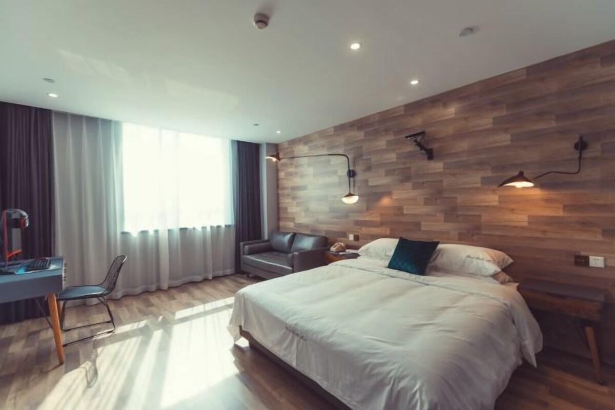 A modern hotel room with a wooden accent wall, a large bed, a desk with a chair, and a window with curtains.