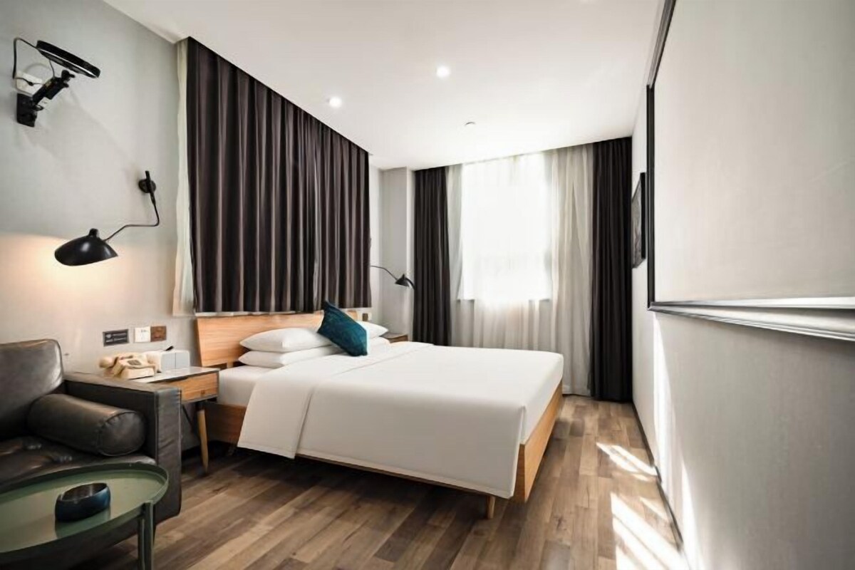 A modern hotel room with a bed, a desk, a chair, a lamp, and a window with curtains.