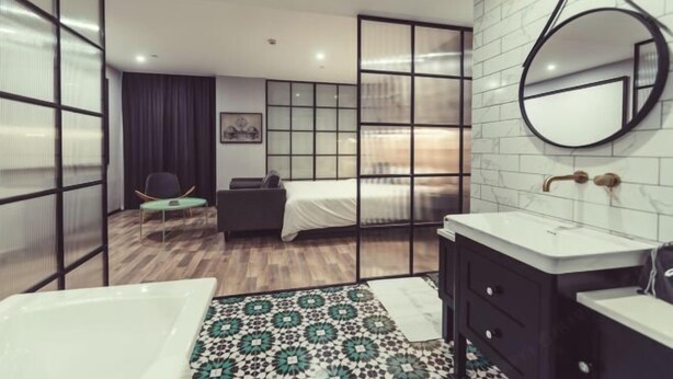 Una habitación de hotel moderna con una cama grande, una zona de estar con una silla, un espejo redondo, una bañera y un lavabo.
