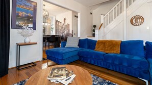 A living room with a blue sectional sofa, a round coffee table with magazines, a framed cityscape artwork, and a staircase leading to another level. - Home Detroit Beautiful Historic  (Detroit)