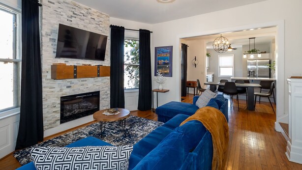 A modern living room with a fireplace, blue sofas, a TV mounted on a brick wall, and a dining area visible through an open doorway. - Home Detroit Beautiful Historic  (Detroit)