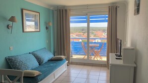 Quarto com parede azul, sofá com almofadas estampadas, uma mesa pequena, televisão e vista para uma cerca e casas azuis pela janela.