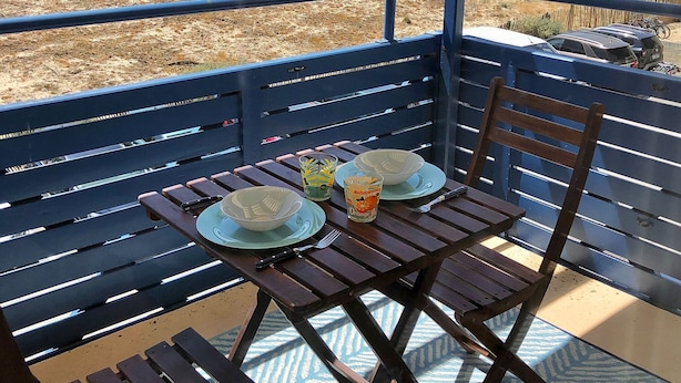 Uma área de estar coberta ao ar livre, com mesa e cadeiras, com vista para uma praia e edifícios ao longe.
