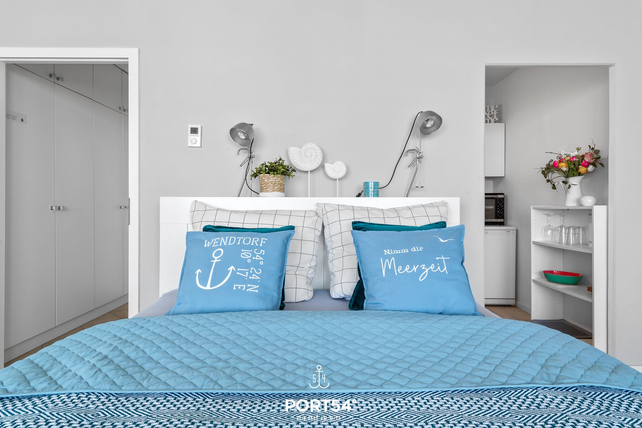 Une chambre à coucher avec un lit, deux oreillers, une table de chevet, un miroir, une lampe fixée au mur et une petite étagère sur laquelle se trouve un vase de fleurs.