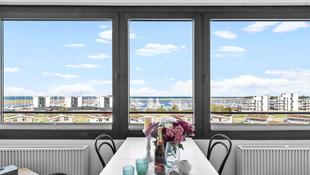 Ein Essbereich mit einem Tisch für zwei Personen, große Fenster mit Blick auf einen Yachthafen und Küstengebäude.