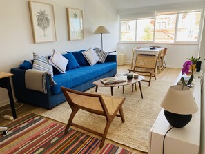 A living room with a blue sofa, a coffee table, a wicker chair, a lamp, and a view of a traditional building through a large window. - Peaceful 2 bd with ocean view in Monte Estoril (Estoril)