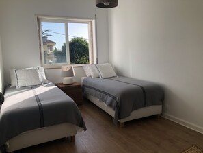 A bedroom with two beds, a window, and a bedside table with a lamp. - Peaceful 2 bd with ocean view in Monte Estoril (Estoril)