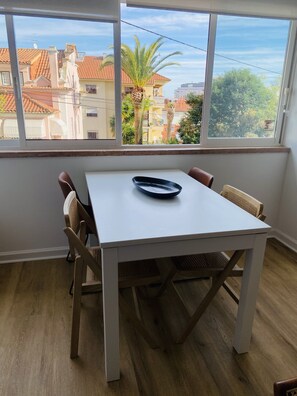 A room with a white table and chairs, a window with a view of buildings and trees, and a black bowl on the table. - Peaceful 2 bd with ocean view in Monte Estoril (Estoril)