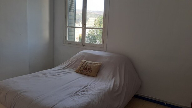 Un lit simple dans une chambre avec une fenêtre, des murs sobres et une porte bien visible.