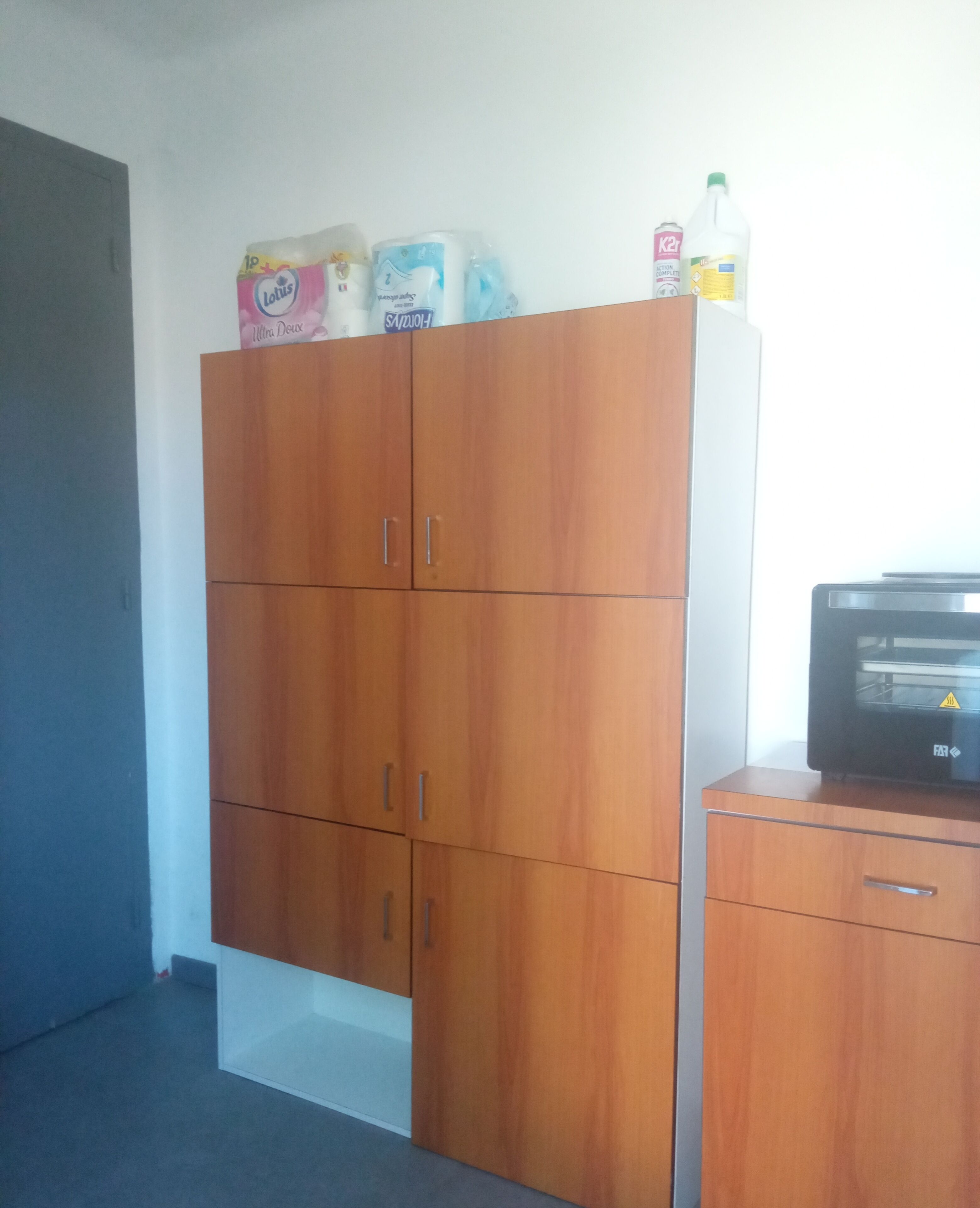 A room with a wooden wardrobe, a small desk with a microwave, and various items on top of the wardrobe including a bottle and a box.