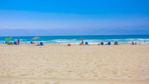 Ein Strand mit Menschen, die unter Sonnenschirmen entspannen, einem klaren Himmel und dem Meer im Hintergrund.