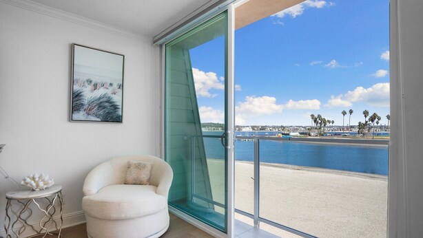 Ein Zimmer mit einer großen Schiebeglasdoor zum Strandblick, ein weißer Sessel, ein kleiner runder Tisch mit einem dekorativen Gegenstand und ein gerahmtes Bild an der Wand.
