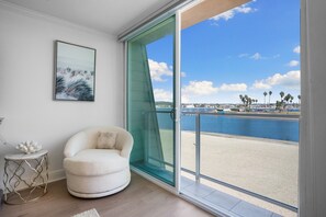 Une pièce dotée d’une grande porte-fenêtre coulissante donnant sur la plage, d’un fauteuil blanc, d’une petite table ronde agrémentée d’un objet décoratif et d’un tableau encadré au mur.