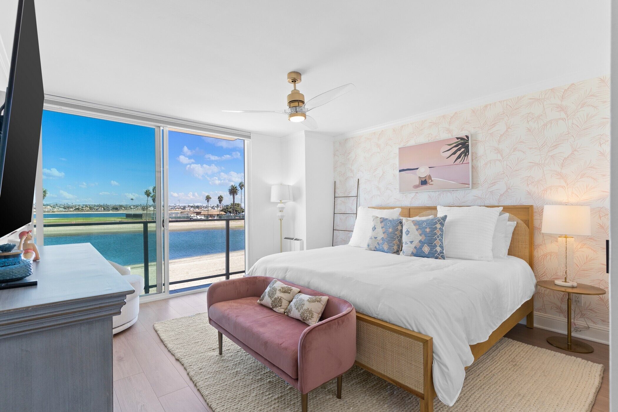 A bedroom with a large bed, a view of the sea, a ceiling fan, and a small table with a lamp.