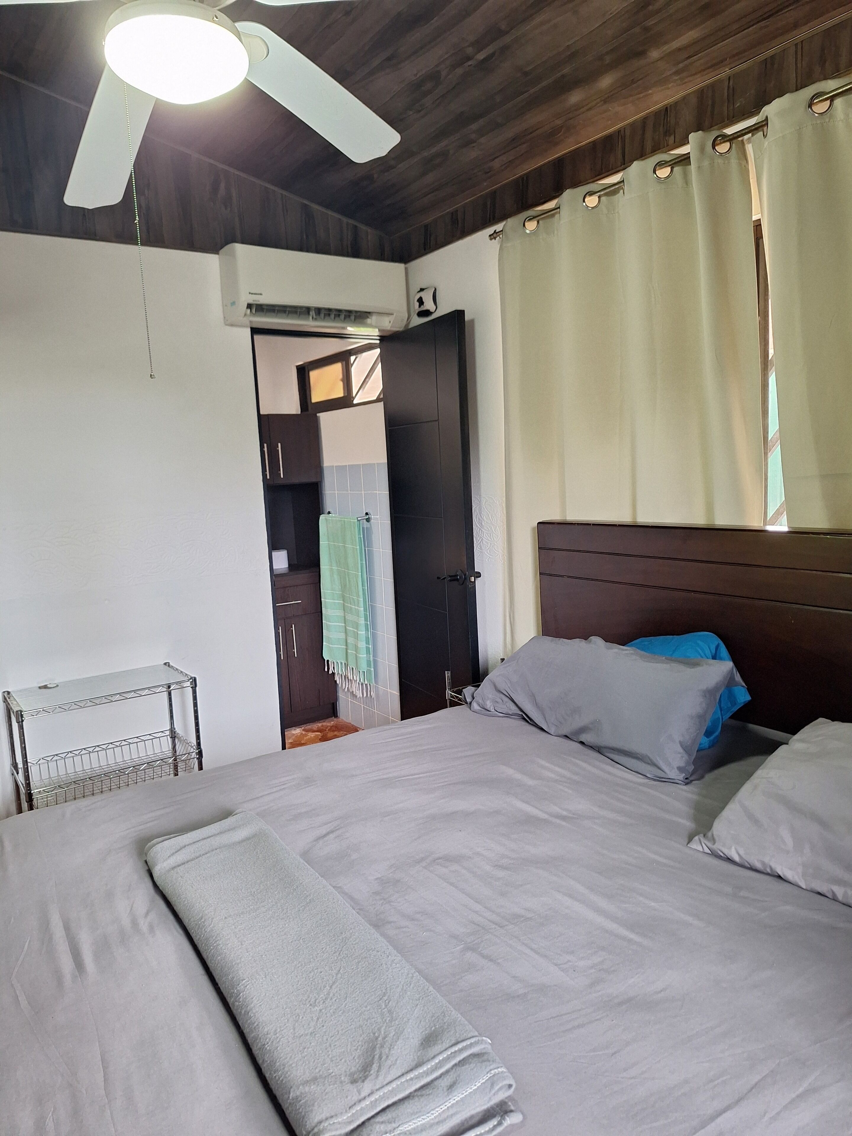Un dormitorio con cama, ventilador de techo, equipo de aire acondicionado, estante metálico y puerta que da a otra habitación.
