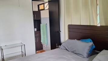 Un dormitorio con cama, ventilador de techo, equipo de aire acondicionado, estante metálico y puerta que da a otra habitación.