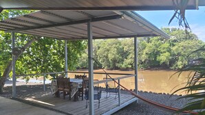 Ein überdachter Außenbereich mit Tisch und Stühlen, einer Hängematte und Blick auf einen Fluss und Wald.