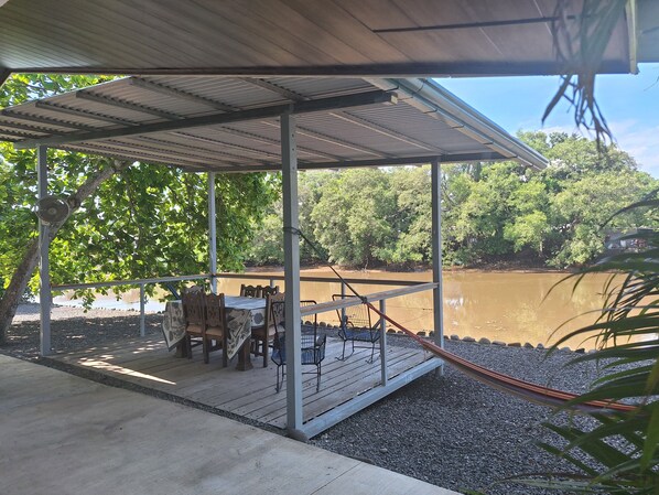 Un espace extérieur abrité, équipé d’une table et de chaises, d’un hamac, et offrant une vue sur une rivière et une forêt.