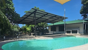 Ein Schwimmbad mit überdachter Terrasse, umgeben von viel Grün und einem Gebäude mit Gründach.