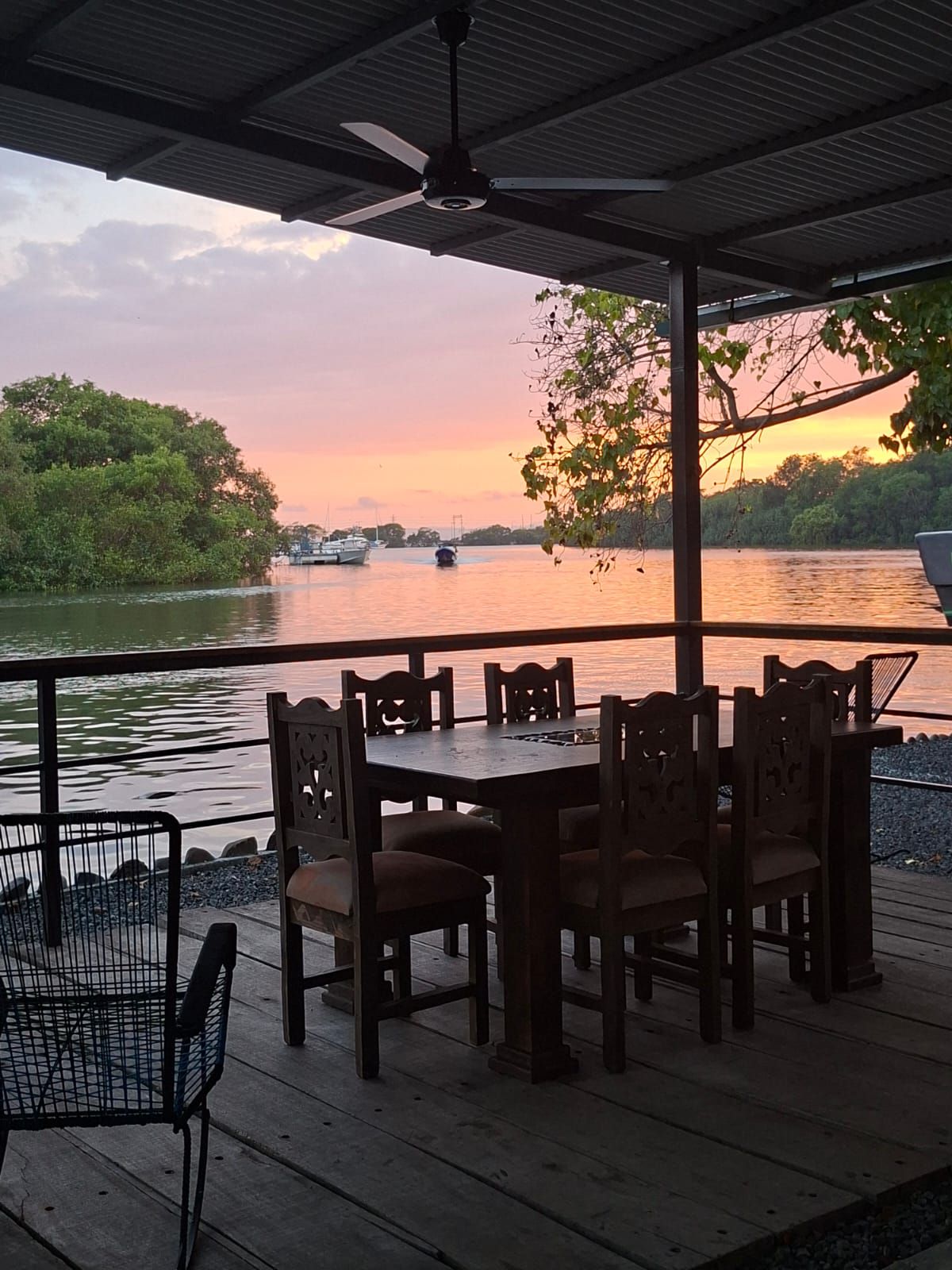 An outdoor dining area with a table and chairs, overlooking a body of water with boats and a sunset in the background.