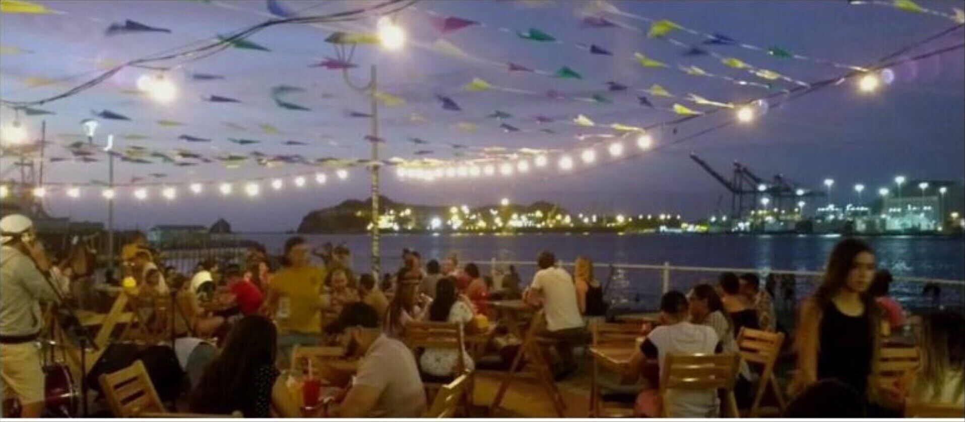A crowded outdoor dining area with tables and chairs, a view of the water, and colorful decorations hanging from the ceiling.