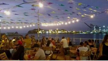 A crowded outdoor dining area with tables and chairs, a view of the water, and colorful decorations hanging from the ceiling.