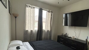 Une chambre à coucher avec un lit, un ventilateur de plafond, un téléviseur fixé au mur et une fenêtre avec des rideaux.