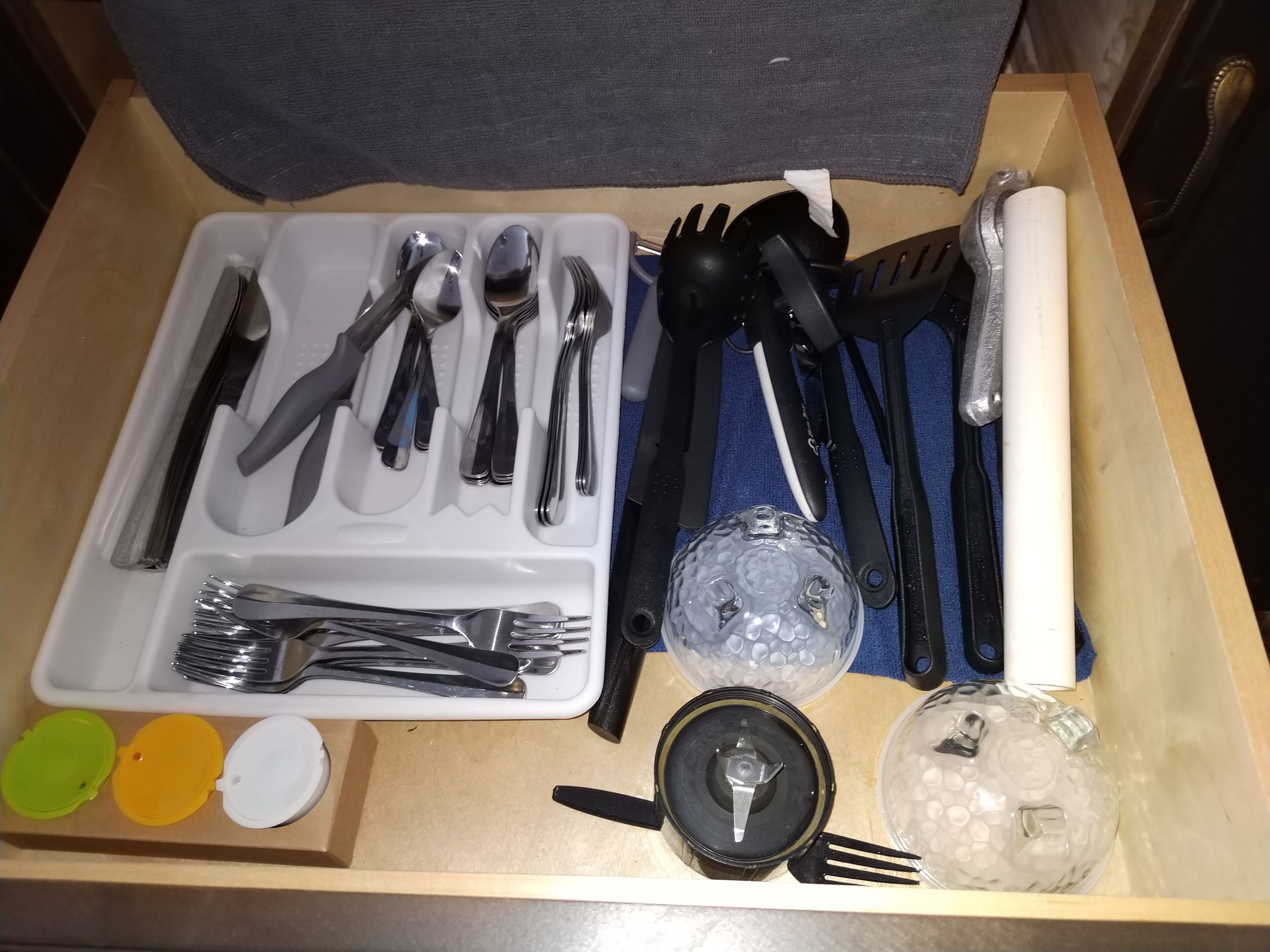 Un calaix amb una selecció de coberts de plata, ganivets i forquilles negres i blancs, una espàtula negra, un rodillo blanc i un petit recipient amb tapa negra.